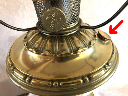 Close-up of a brass antique lamp base with an ornate pattern and a red arrow pointing to a small knob.