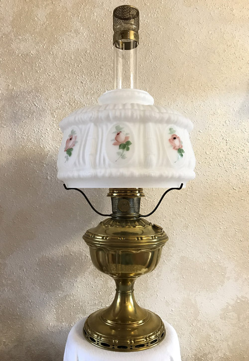 Vintage brass oil lamp with white glass shade decorated with pink roses on a textured wall background.