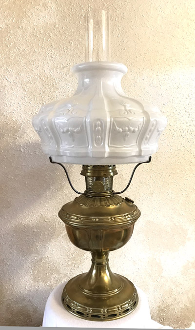 Antique brass oil lamp with ornate white glass shade and clear chimney on textured wall.