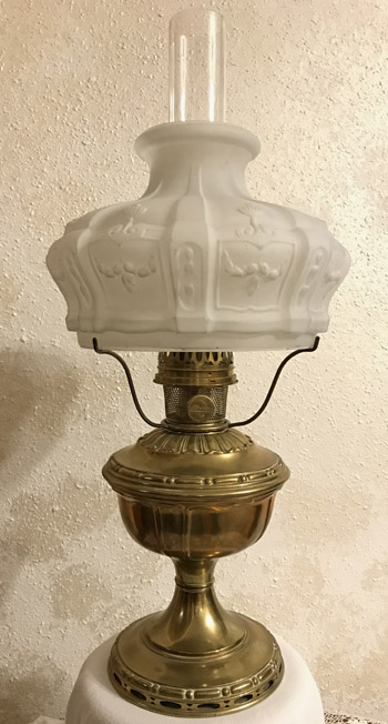 Antique brass oil lamp with a frosted glass shade and clear glass chimney on a textured wall.