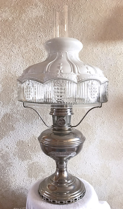 Vintage silver oil lamp with a clear glass midsection and white frosted glass shade on textured wall.