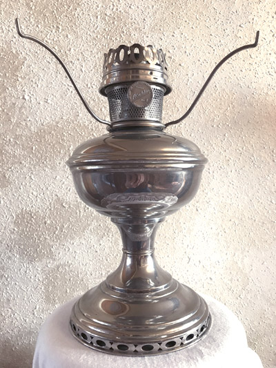 Shiny metal antique oil lamp with handle arms on textured beige wall background.