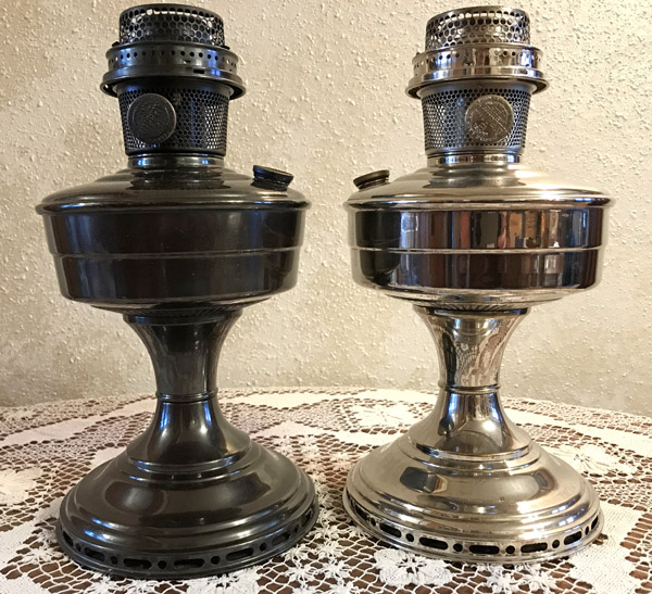 Two vintage metal oil lamps, one dark and one shiny, on a lace tablecloth against a beige wall.