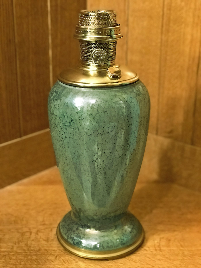 Green ceramic vase with a brass oil lamp burner on top, placed on a wooden surface and background.