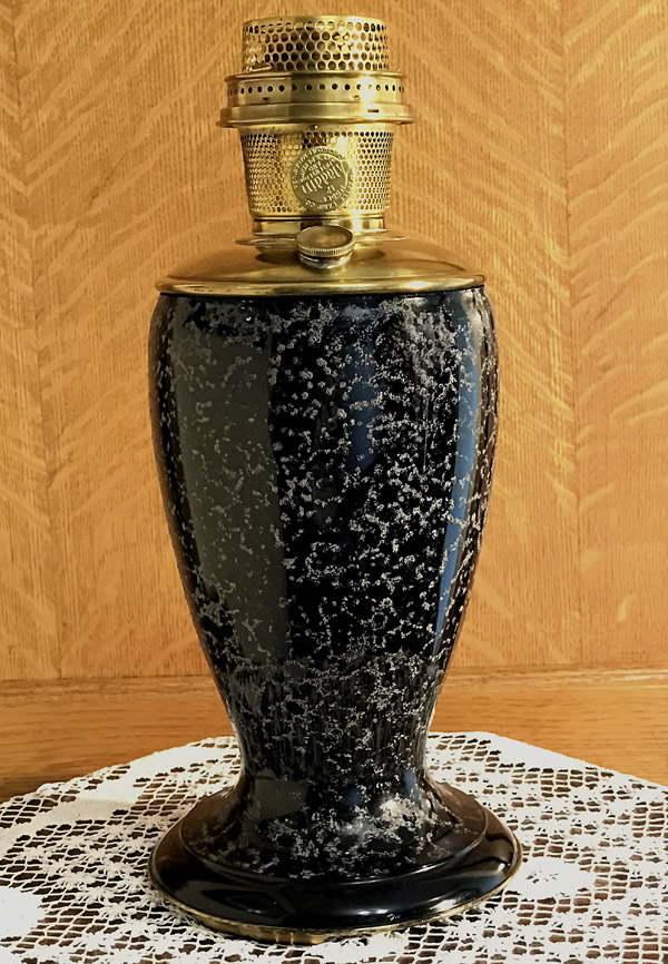 Tall black and gray speckled vintage oil lamp with brass burner on lace doily and wooden background.