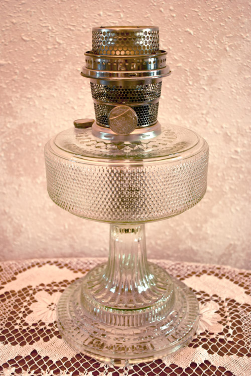 Vintage clear glass oil lamp with metal burner sitting on a lace tablecloth against a textured wall.