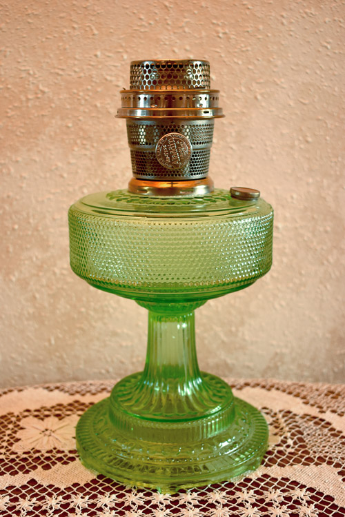 Green vintage glass oil lamp with metal burner on lace tablecloth against textured wall.