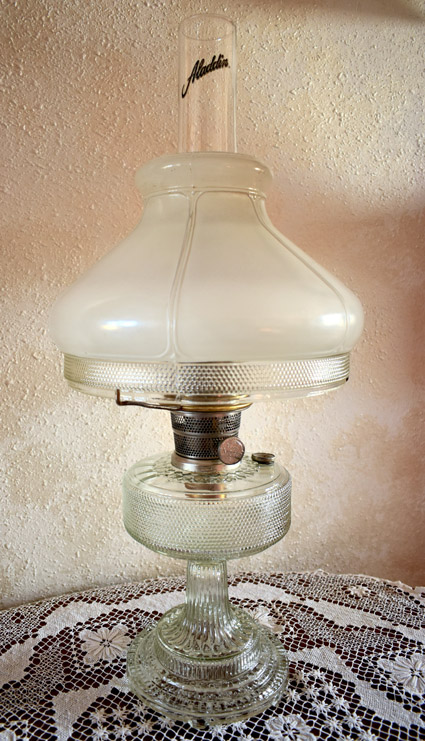 Vintage glass Aladdin oil lamp with frosted chimney and ornate base on lace tablecloth.