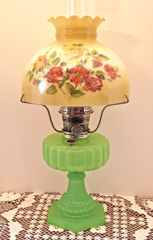 Vintage green glass oil lamp with floral-patterned cream glass shade on a lace tablecloth.