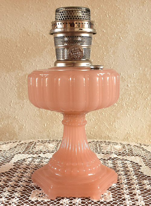 Vintage pink glass oil lamp with ornate base on a lace cloth background.