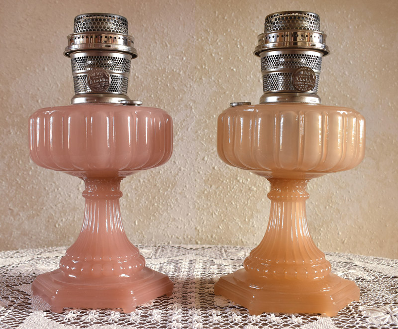 Two vintage glass oil lamps with metal burners, one pink and one orange, on a lace tablecloth.