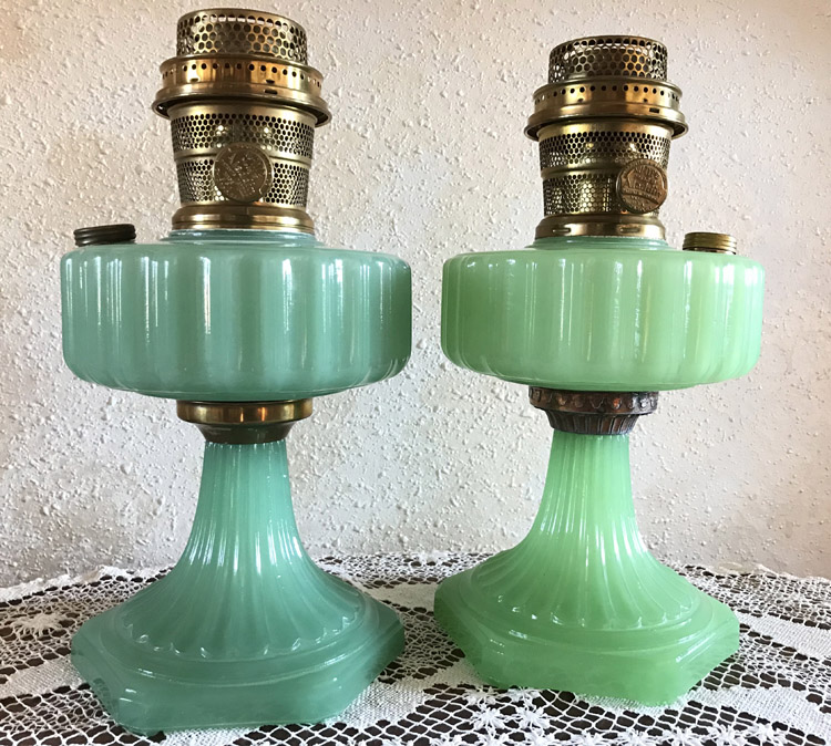 Two vintage green glass hurricane lamps with brass tops on a lace-covered surface.