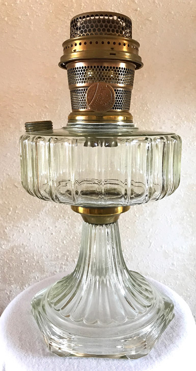 Vintage clear glass oil lamp with ribbed base and brass burner against beige wall.