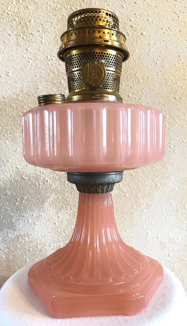 Antique pink glass oil lamp with ribbed design and brass burner on textured wall background.