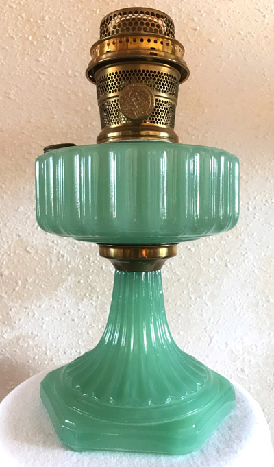 Vintage jade green glass Corinthian oil lamp with brass burner on a white surface and textured wall background.