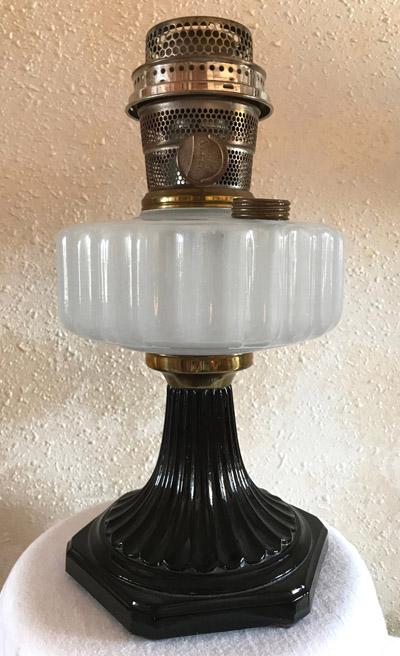 Vintage oil lamp with a black ornate base, frosted ribbed glass reservoir, and a metal burner on top.