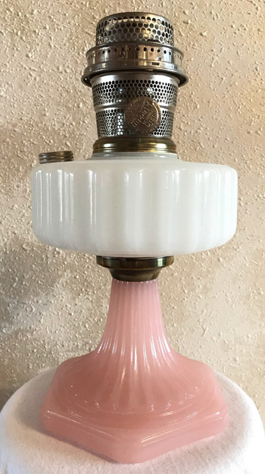 Vintage oil lamp with white ribbed glass reservoir and pink ribbed glass base against textured beige wall.