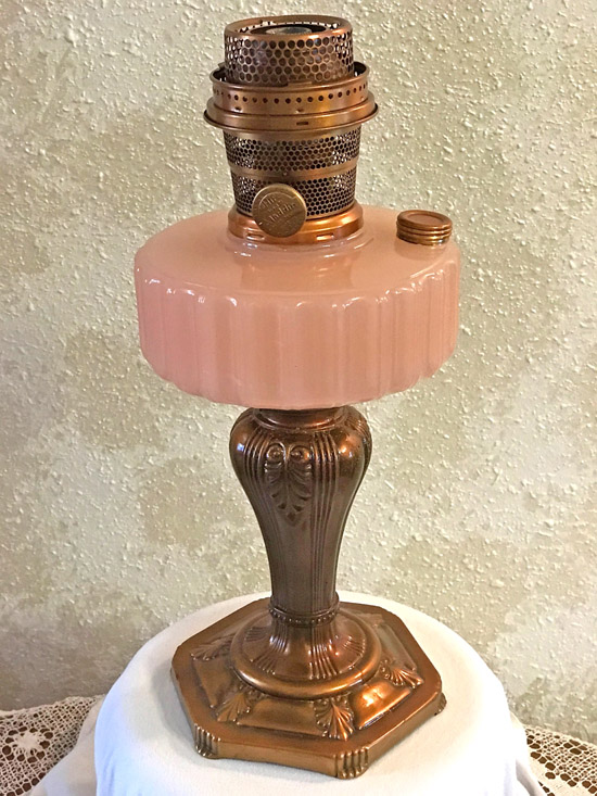Vintage oil lamp with rose pink glass reservoir and ornate brass base on white cloth.