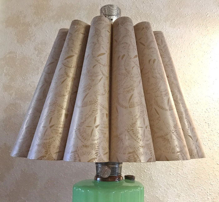 Beige fluted lamp shade with subtle pattern on a green lamp base against textured wall.