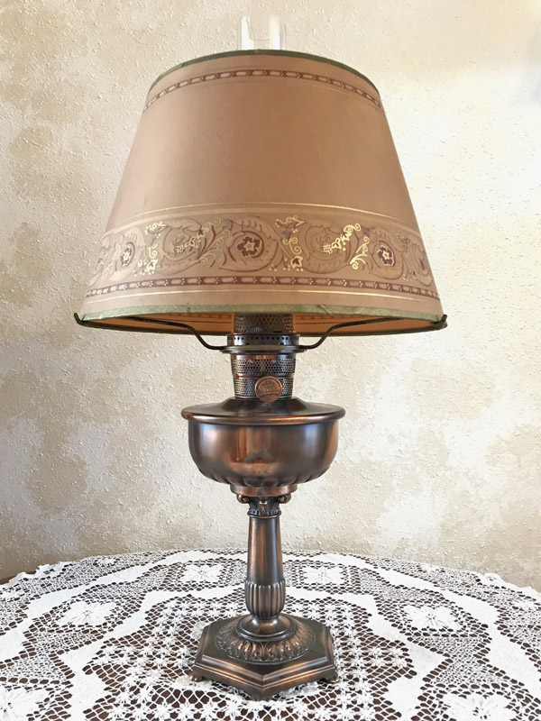 Vintage rose gold table lamp with ornate base and beige lampshade with gold accents on lace cloth.
