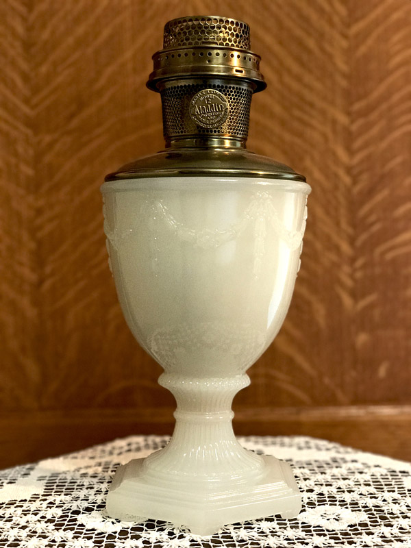 Ornate white milk glass oil lamp with brass burner on lace cloth against wooden background.