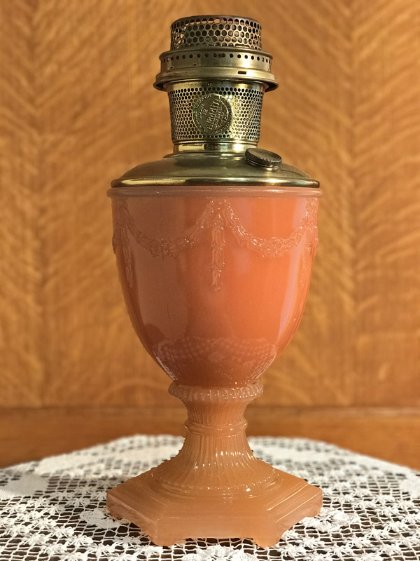 Vintage pink glass lamp with brass burner on a lace-covered wooden surface.