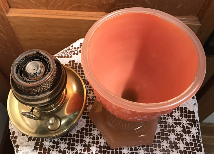 Top view of a vintage oil lamp with a brass burner and an orange glass chimney on a lace tablecloth.