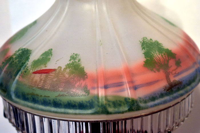 Close-up of a lampshade with a painted landscape featuring trees, a cabin, and a red sunset sky.