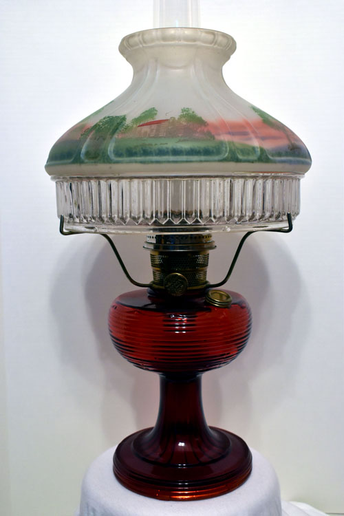 Vintage red glass oil lamp with decorative glass shade featuring a painted rural scene.
