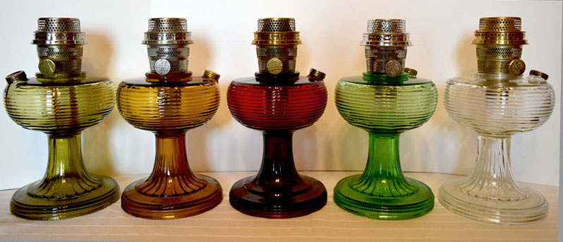 Five vintage glass oil lamps in different colors arranged in a row on a white surface.