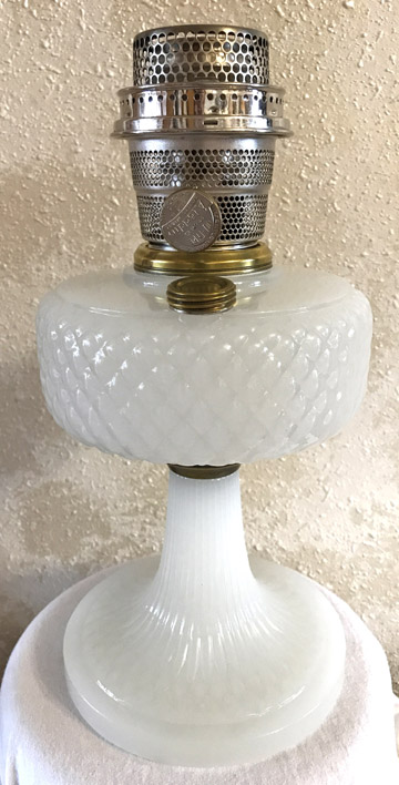 Vintage white glass oil lamp with textured reservoir and metal burner on a textured beige background.