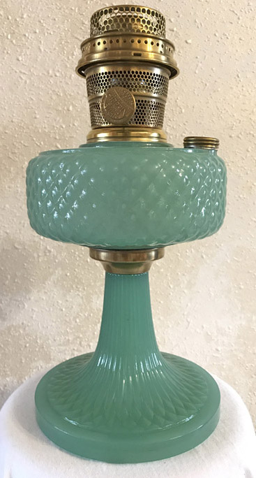Vintage green glass oil lamp with brass burner and a textured base against white wall.