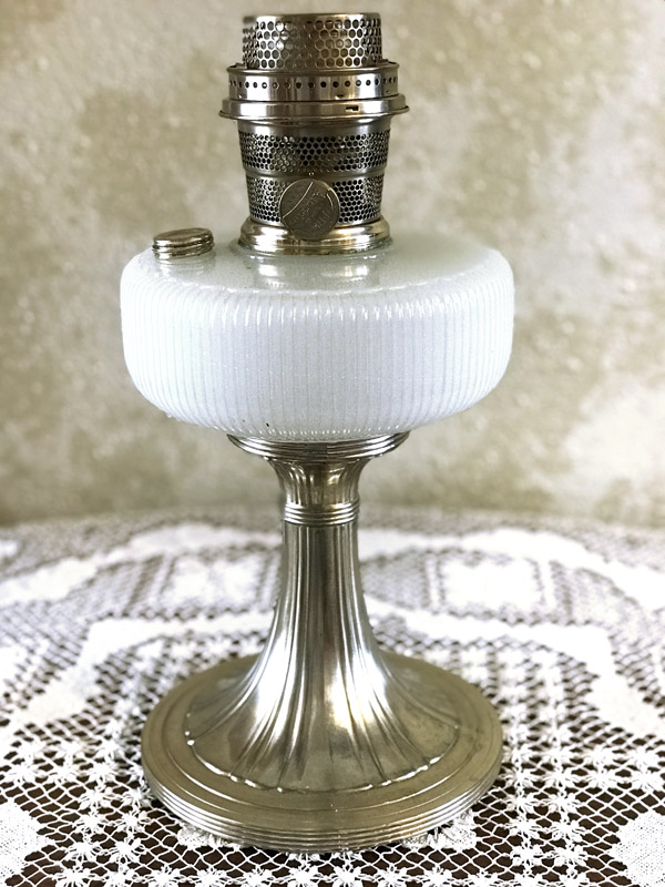 Vintage metal and white glass oil lamp on a lace-patterned cloth surface.