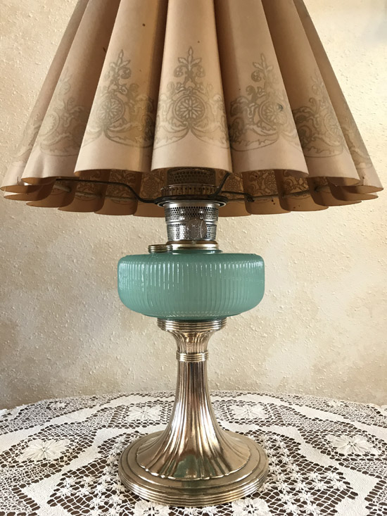 Vintage table lamp with a pleated beige shade, aqua glass reservoir, and silver metal base on lace tablecloth.