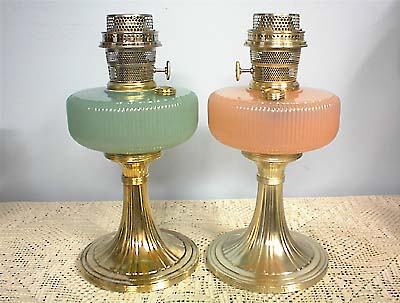 Two vintage glass oil lamps with brass bases, one green and one pink, on a lace tablecloth.