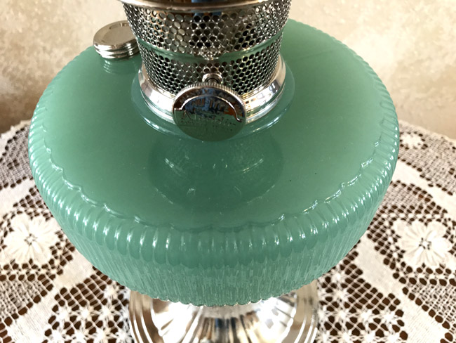 Close-up of a vintage green glass lamp with metal burner on a lace-covered table.