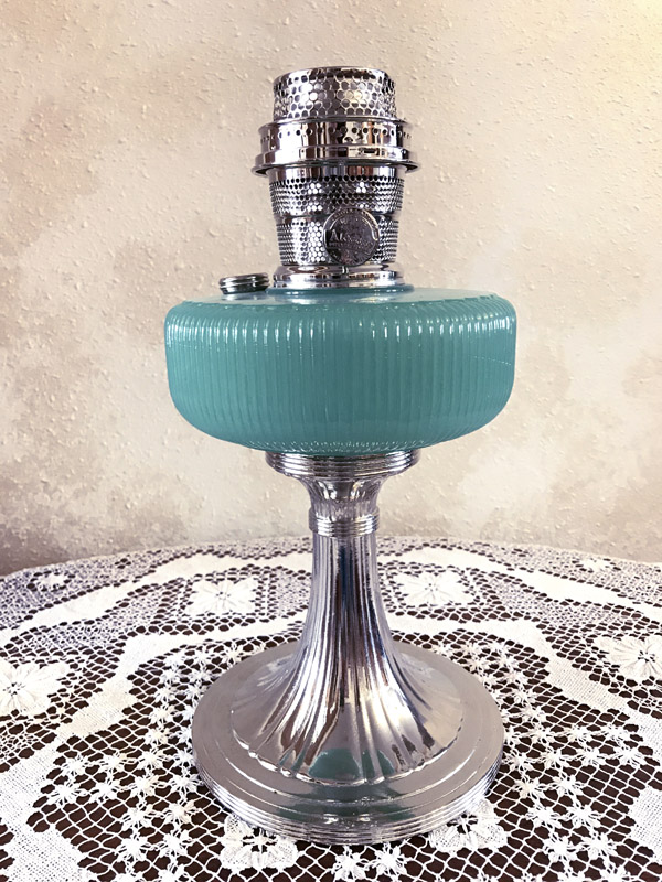 Vintage silver oil lamp with a turquoise glass font on a lace-covered surface.