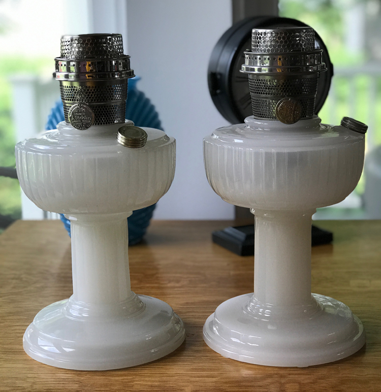Pair of white vintage Aladdin oil lamps with metal tops on a wooden surface.