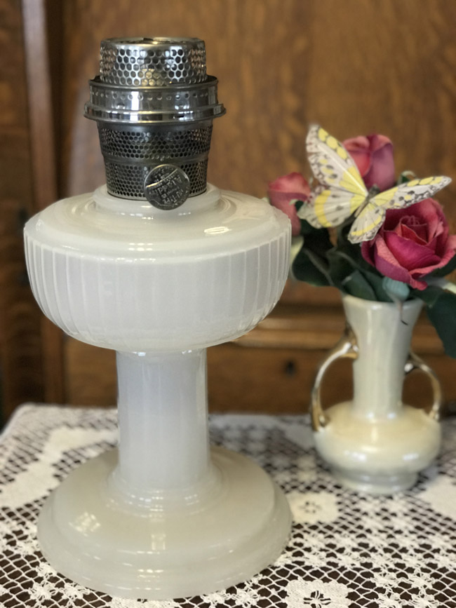 White vintage oil lamp on lace tablecloth beside vase with pink flowers and yellow butterfly decoration.