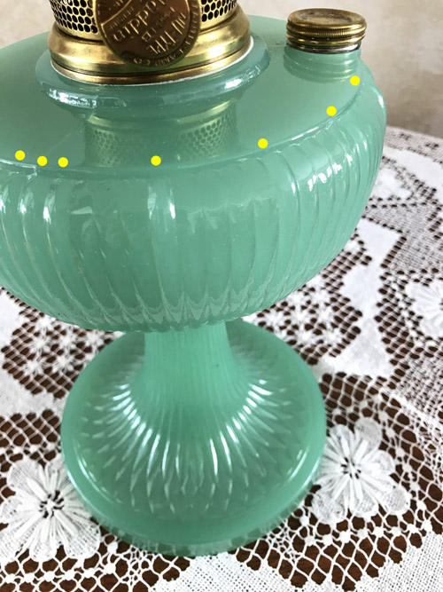 Close-up of a green ribbed glass oil lamp with brass burner on a lace tablecloth.