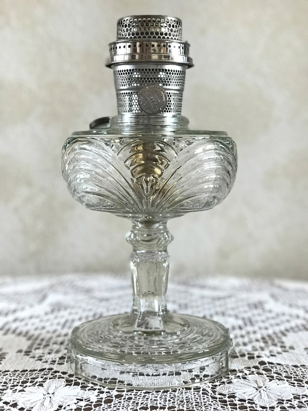 Clear glass oil lamp with ornate filigree stem and metal burner on lace cloth.