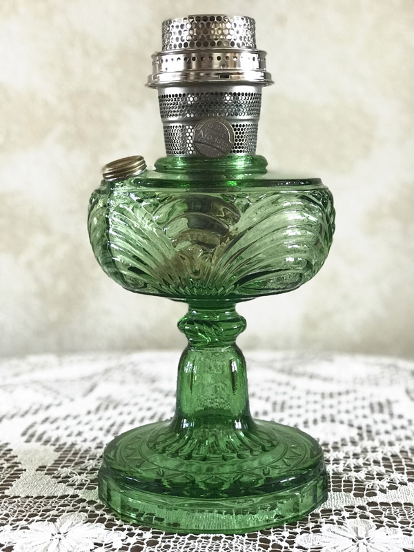 Green glass oil lamp with decorative patterns on a lace tablecloth against a neutral background.
