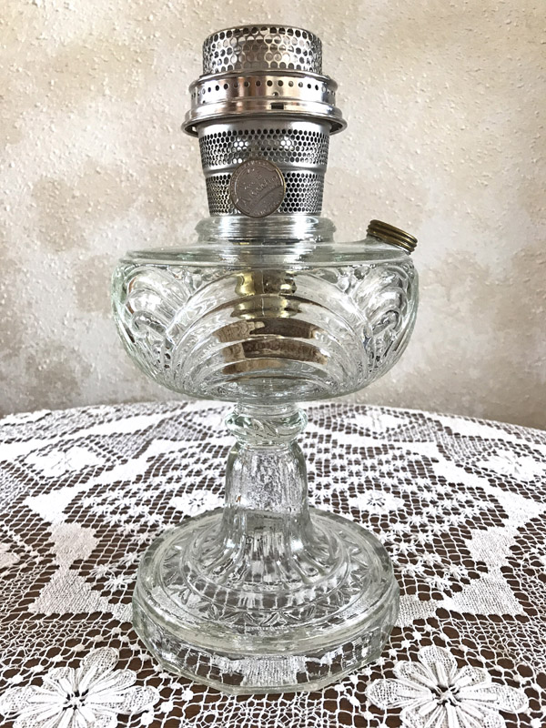 Clear glass oil lamp with ornate stem and metal burner on a white lace tablecloth.