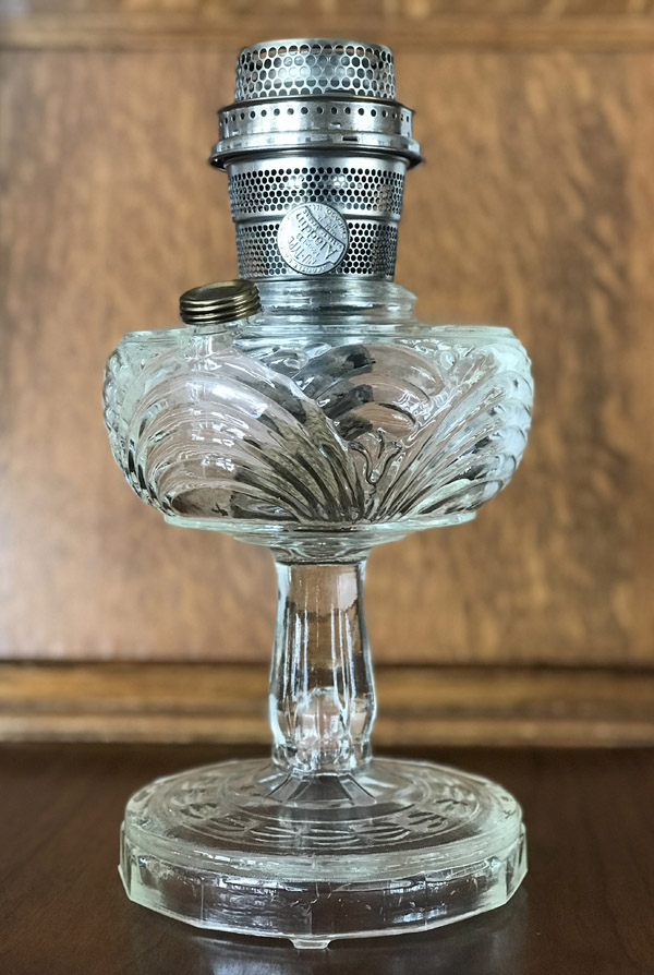 Antique clear glass oil lamp with ornate patterns and metal burner on wooden surface.