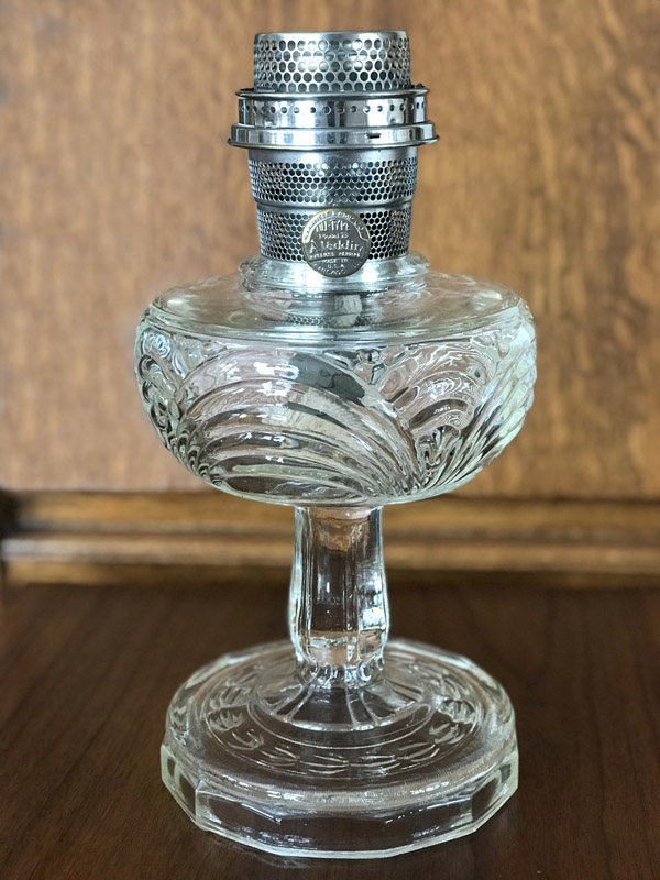 Clear glass oil lamp with ornate base and metal burner on a wooden surface and background.