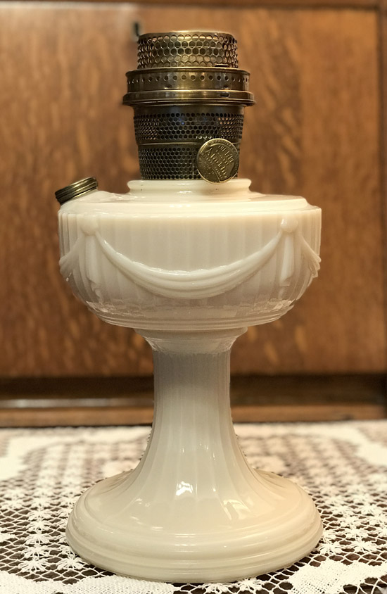 White vintage oil lamp with decorative draped design and metal burner on a lace tablecloth.