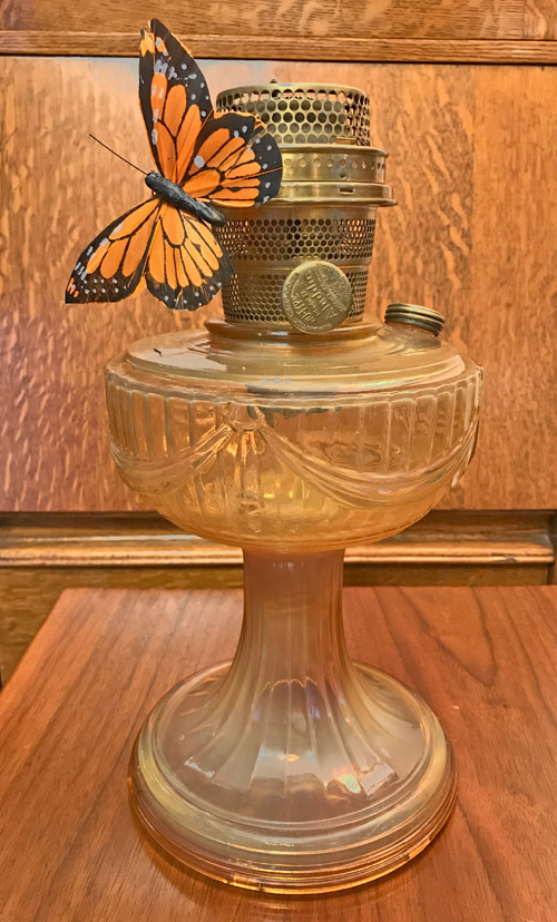 Orange and black butterfly perched on the metal top of an amber glass oil lamp on a wooden surface.