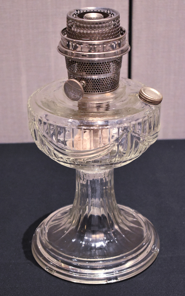 Antique clear glass oil lamp with metal burner and wick adjuster on a pedestal base.