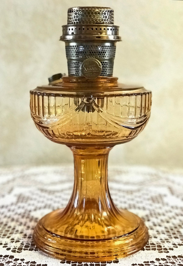 Amber-colored vintage glass oil lamp with metal burner on a white lace tablecloth.