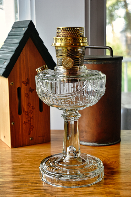 Antique glass oil lamp with brass burner on wooden table, beside a wooden birdhouse and a metal container.